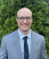 a bald man in glasses and a suit smiles in front of bushes